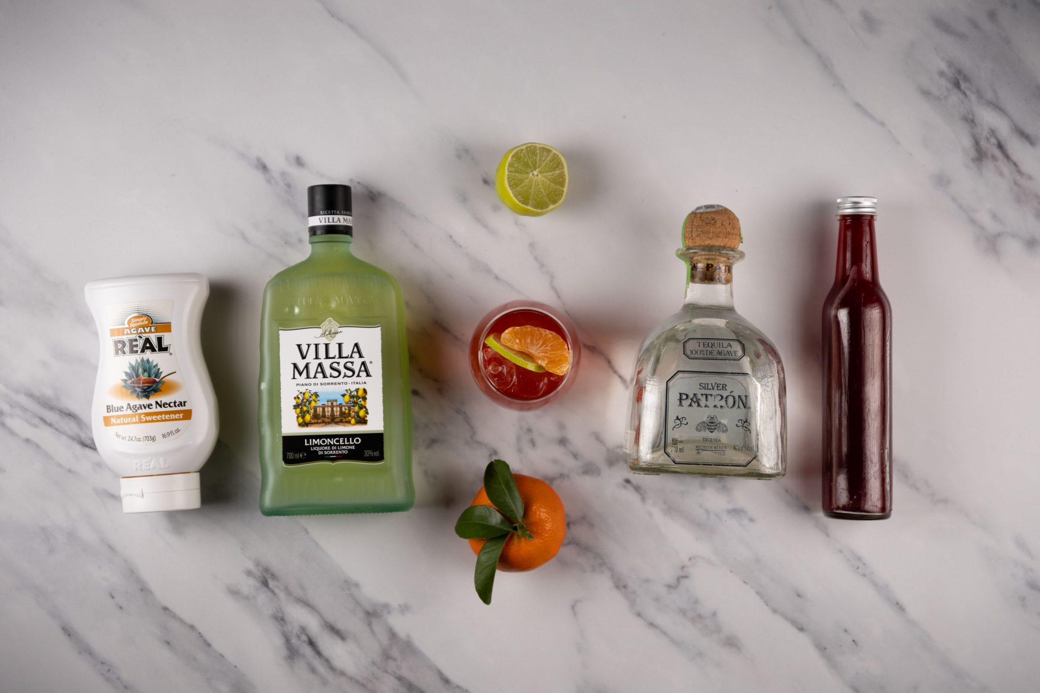 A Holiday Margarita cocktail, shot from above, in a rocks glass on a white marmol surface surrounded by Tequila, limoncello, pomegranate juice, lime juice, lime, tangerine juice and agave syrup