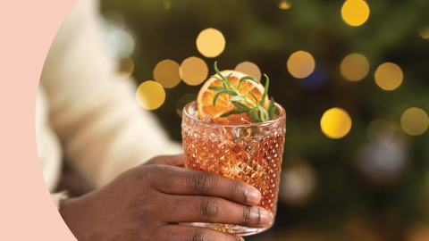 A shot of a hand holding an Old Fashioned glass with an orange cocktail garnished with rosemary sprig and an orange slice.