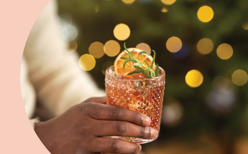 A shot of a hand holding an Old Fashioned glass with an orange cocktail garnished with rosemary sprig and an orange slice.