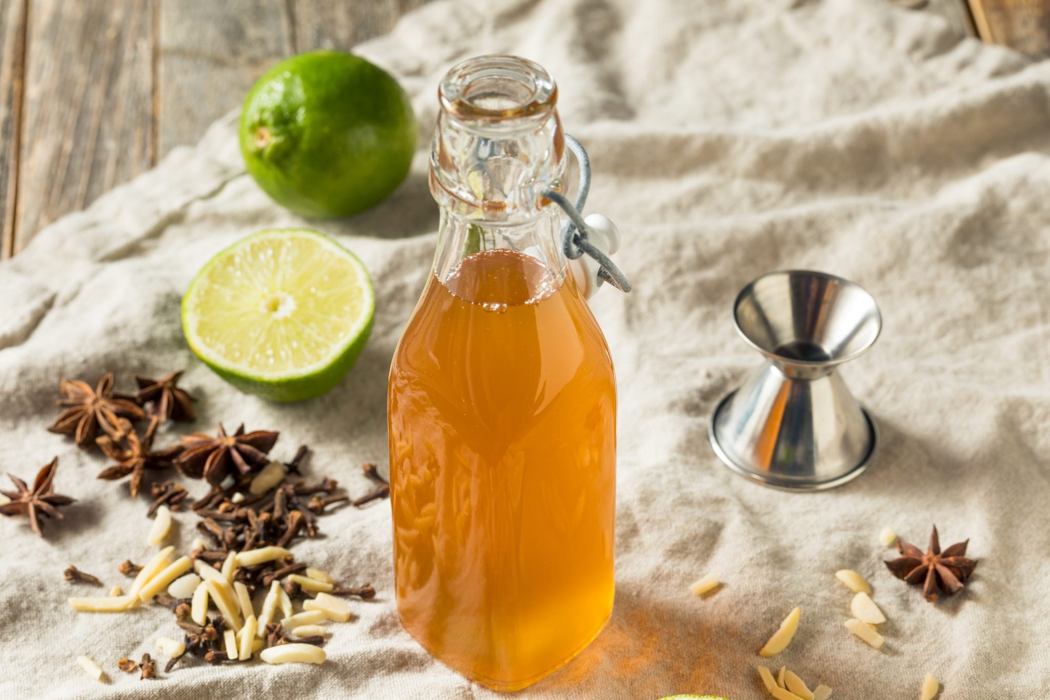Small bottle with homemade Falernum on a white cloth surrounded by two limes, a jigger, star anises, gloves and almonds
