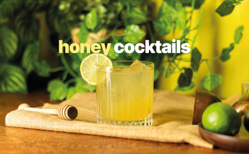 A shot of a yellow cocktail in an Old Fashioned glass on a yellow cloth placed on a wooden table surrounded by a jigger, a lime, and a honey dipper, in front of a yellow background and plant leaves