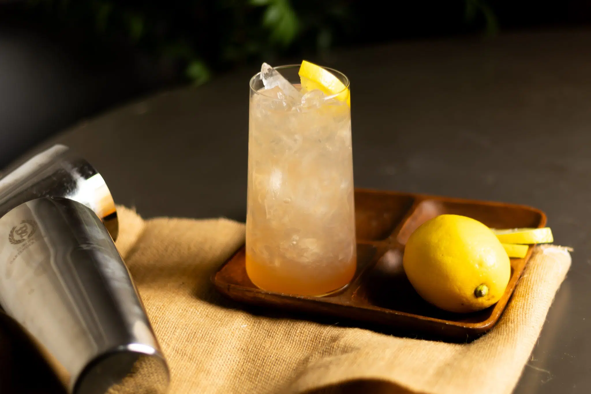 A side shot of a Honey Deuce cocktail in highball glass on a wooden tray placed on a dark grey surface surrounded by an orange cloth, a lemon, two lemon slices, and a shaker, in front of a black background. 