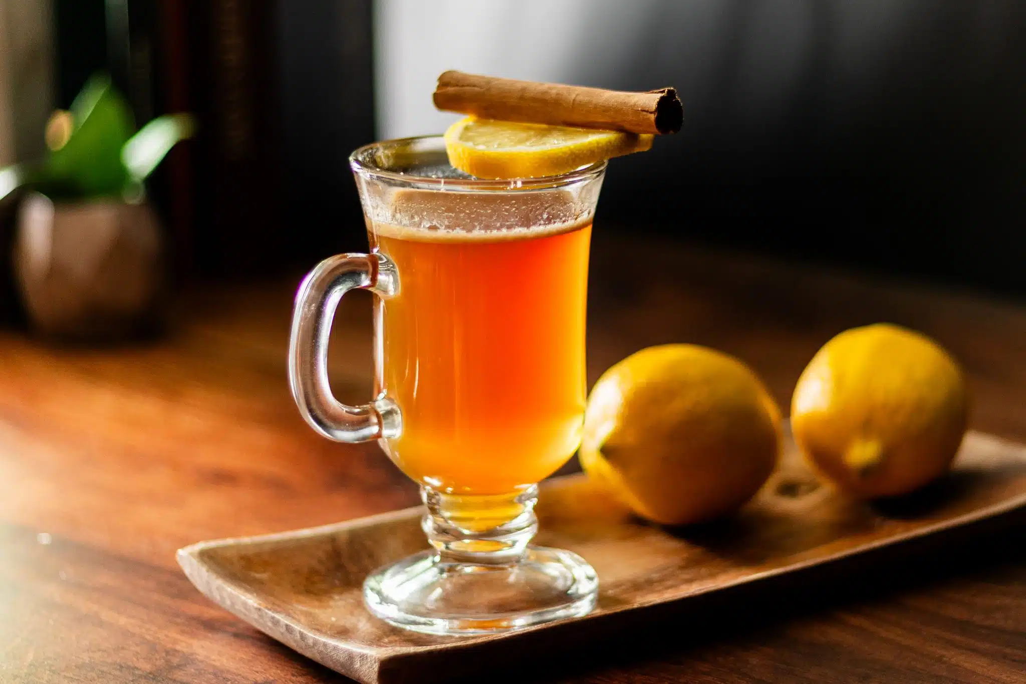 A side shot of a Hot Buttered Rum cocktail in a mug with two lemons behind on a wooden tray placed on a brown wooden table and a plant on the background.