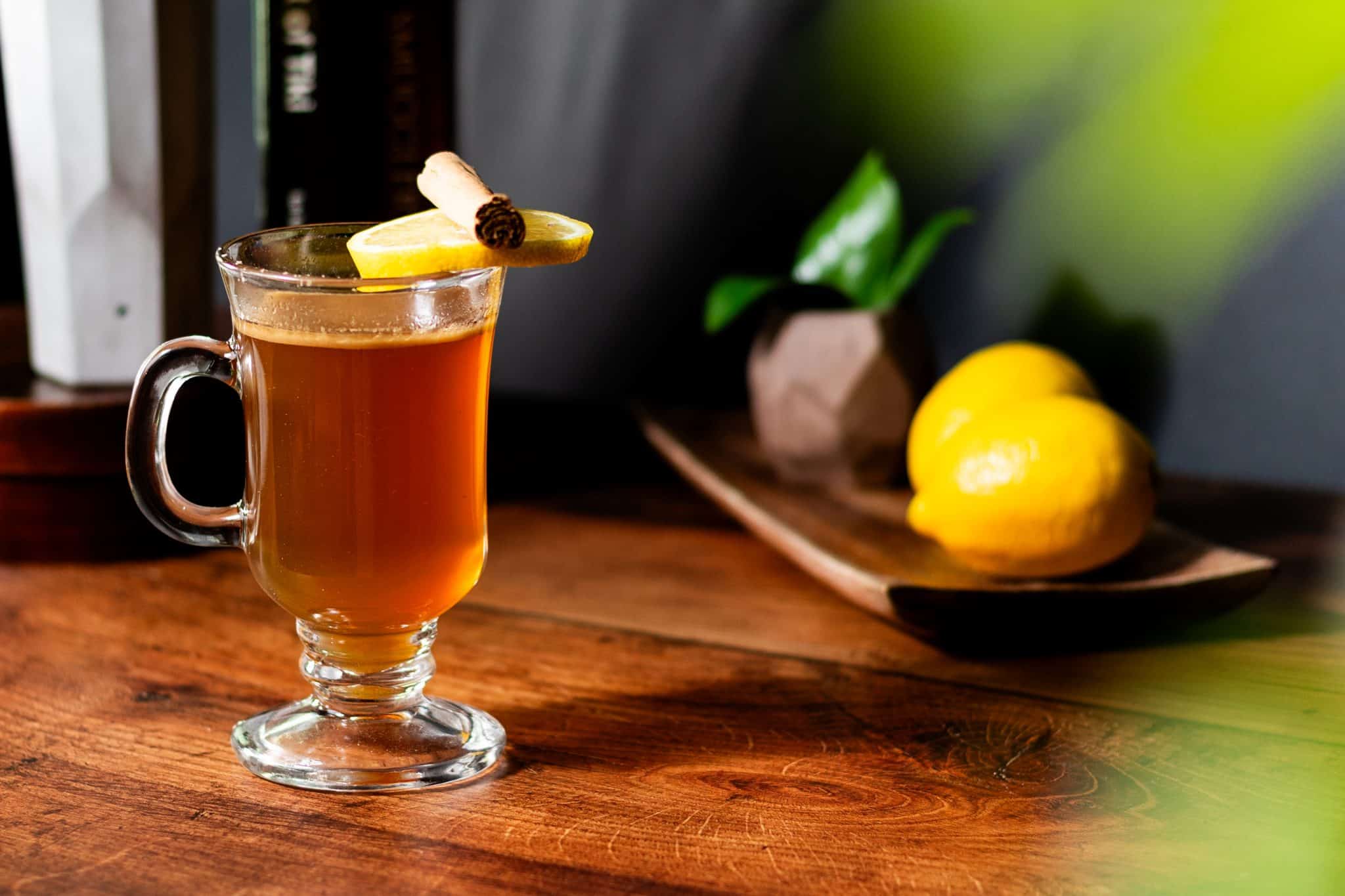 A side shot of a Hot Buttered Rum cocktail in a mug on a brown wooden table with two lemons and a small plant behind on a wooden tray.