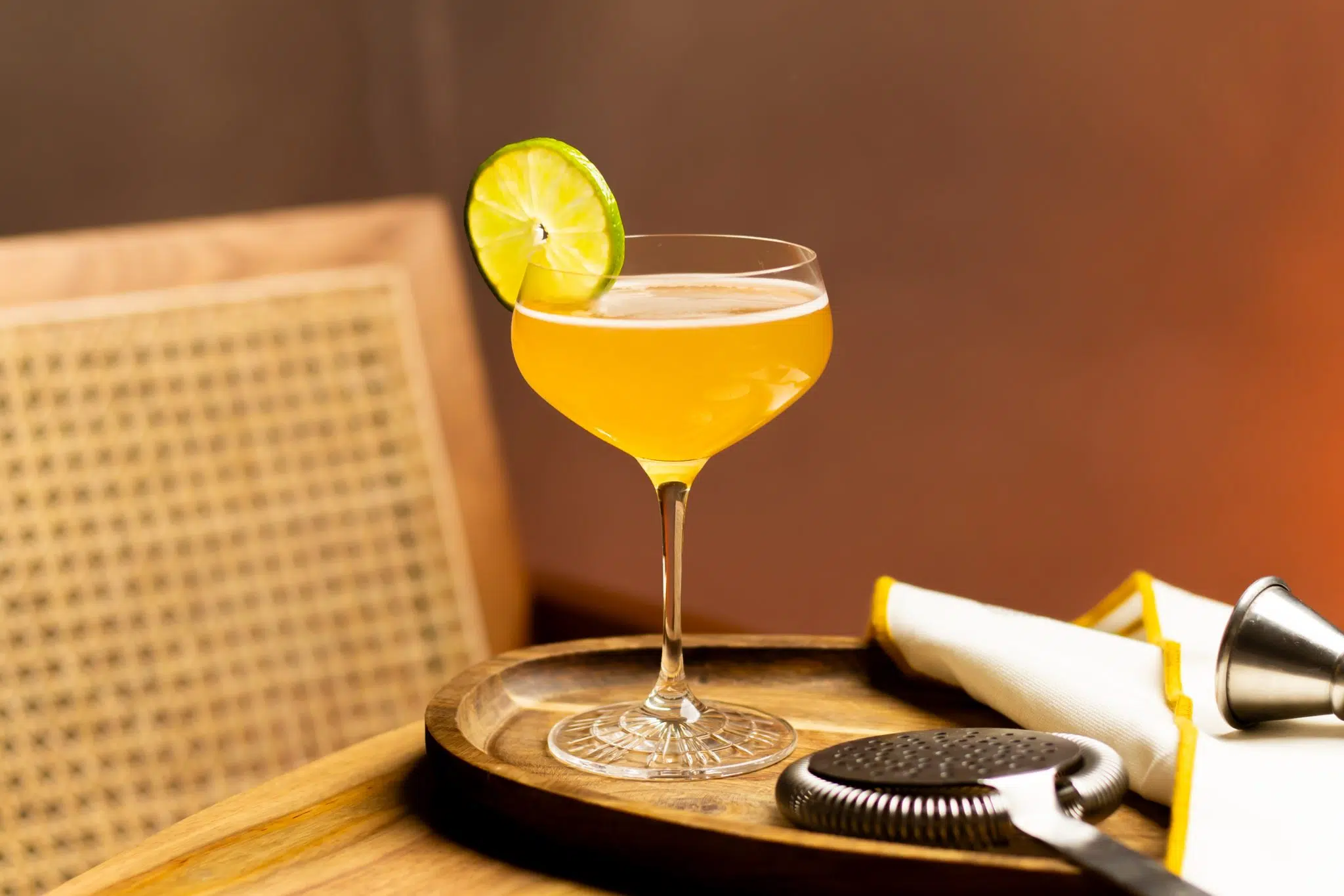 A side shot of a Hotel Nacional Special cocktail in a cocktail glass on a wooden tray with a cocktail strainer, a jigger and a white cloth around.