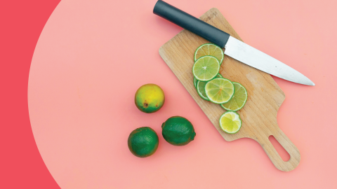 A shot from above of a knife, a wooden board, three limes and limes slices on a pink surface