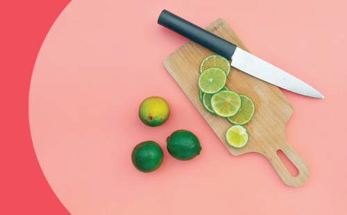 A shot from above of a knife, a wooden board, three limes and limes slices on a pink surface