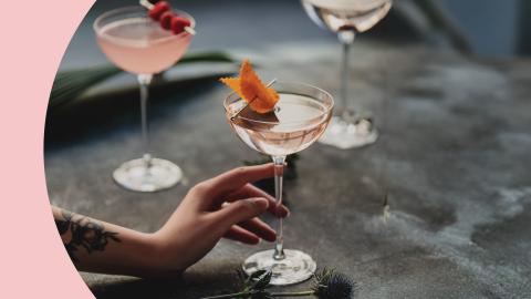 A shot of three cocktail in coupe glasses on a dark grey surface with a hand holding on of them