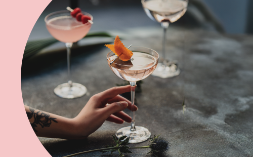 A shot of three cocktail in coupe glasses on a dark grey surface with a hand holding on of them