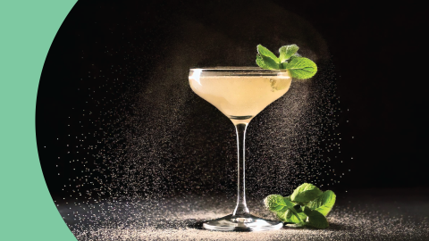 A shot of a cocktail in a coupe glass on a black background with a mint sprig on the side and dust around