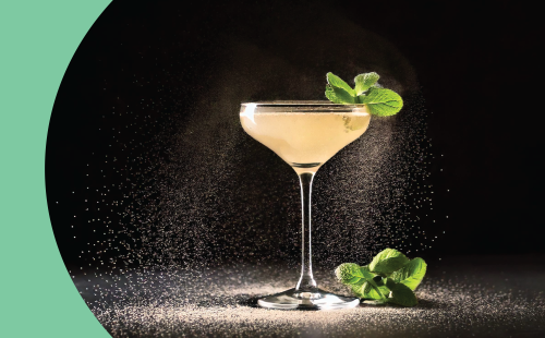 A shot of a cocktail in a coupe glass on a black background with a mint sprig on the side and dust around