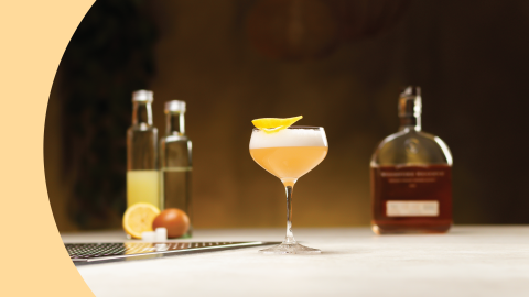 A side shot of a sour cocktail in a coupe glass on a white bar table with a spirit bottle, an orange juice bottle and a simple syrup bottle behind