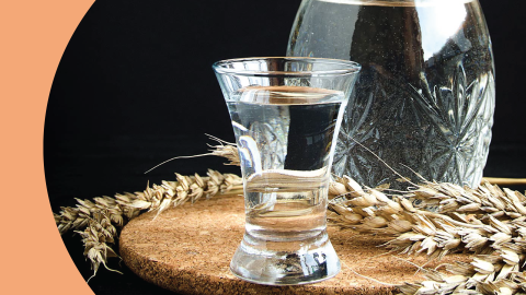 A shot of a vodka shot glass on a cork coaster with a vodka bottle and wheat sprigs in front of a black background