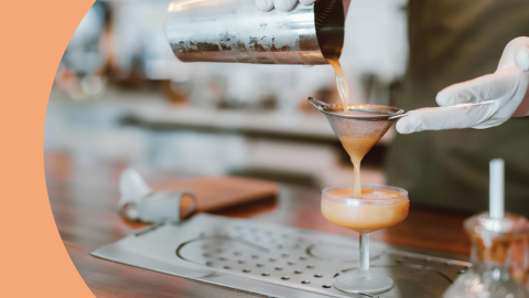 A shot of a bartender with white gloves straining a cocktail in a coupe glass on a bar table