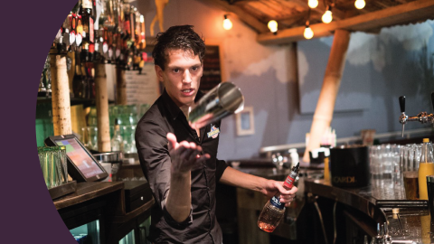 A shot of a bartender throwing a shaker glass behind a bar table