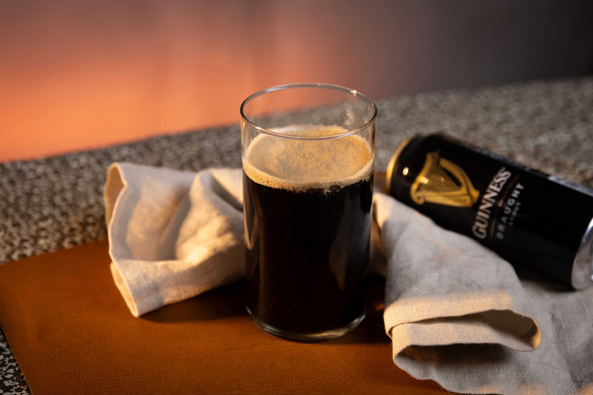 A side shot of a Irish Car Bomb cocktail in a Pint and a shot glass on a brown surface surrounded by a white cloth, and a Guinness beer