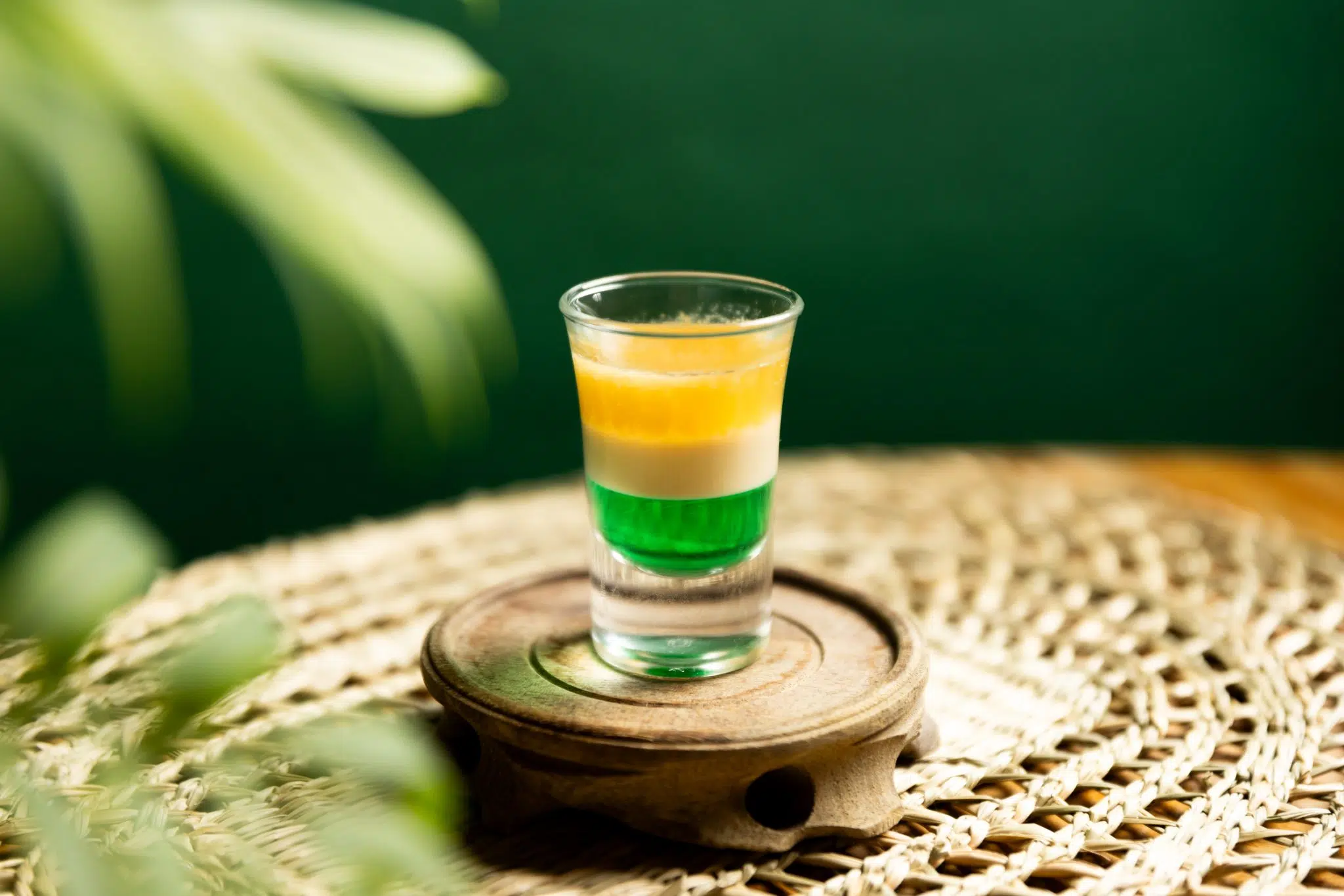 A side shot of a Irish Flag shot in a shot glass on a wooden coaster on a embroidered placemat placed on a brown surface, in front of a green background