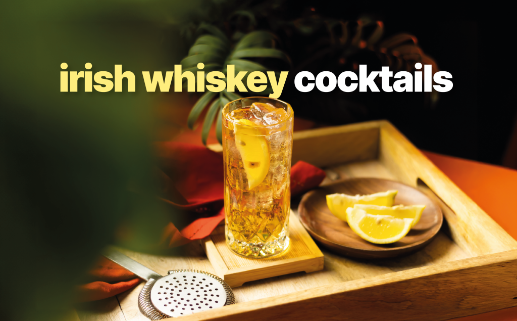 A side shot of an Irish Whiskey cocktail in a highball glass on a wooden coaster and tray surrounded by a cocktail strainer, lemon wedges on a plate and a red cloth