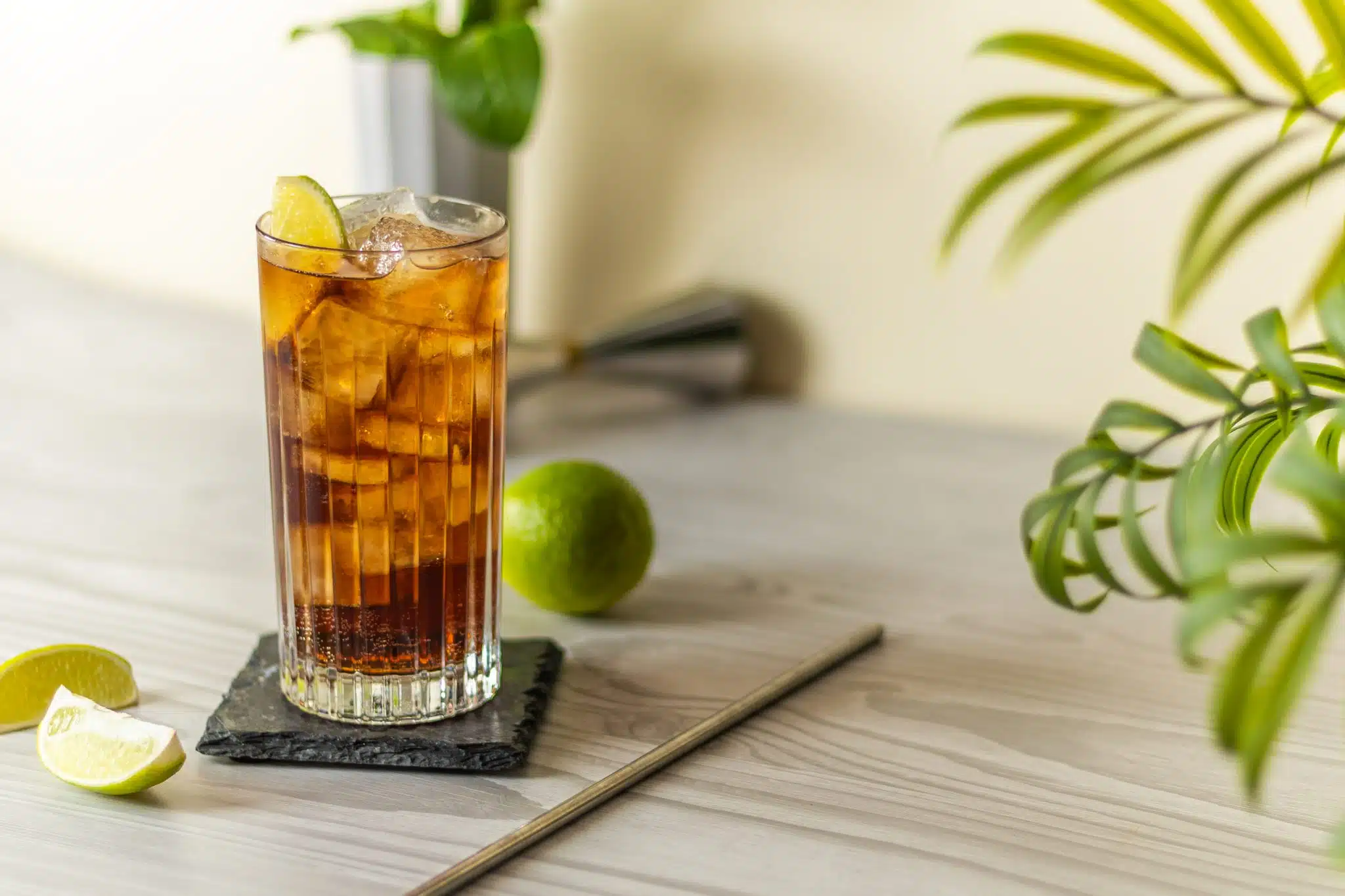 A side shot of a Jack and Coke cocktail in a highball glass on a black stone plate placed on a light grey table surrounded by a straw, two lime wedges and a lime