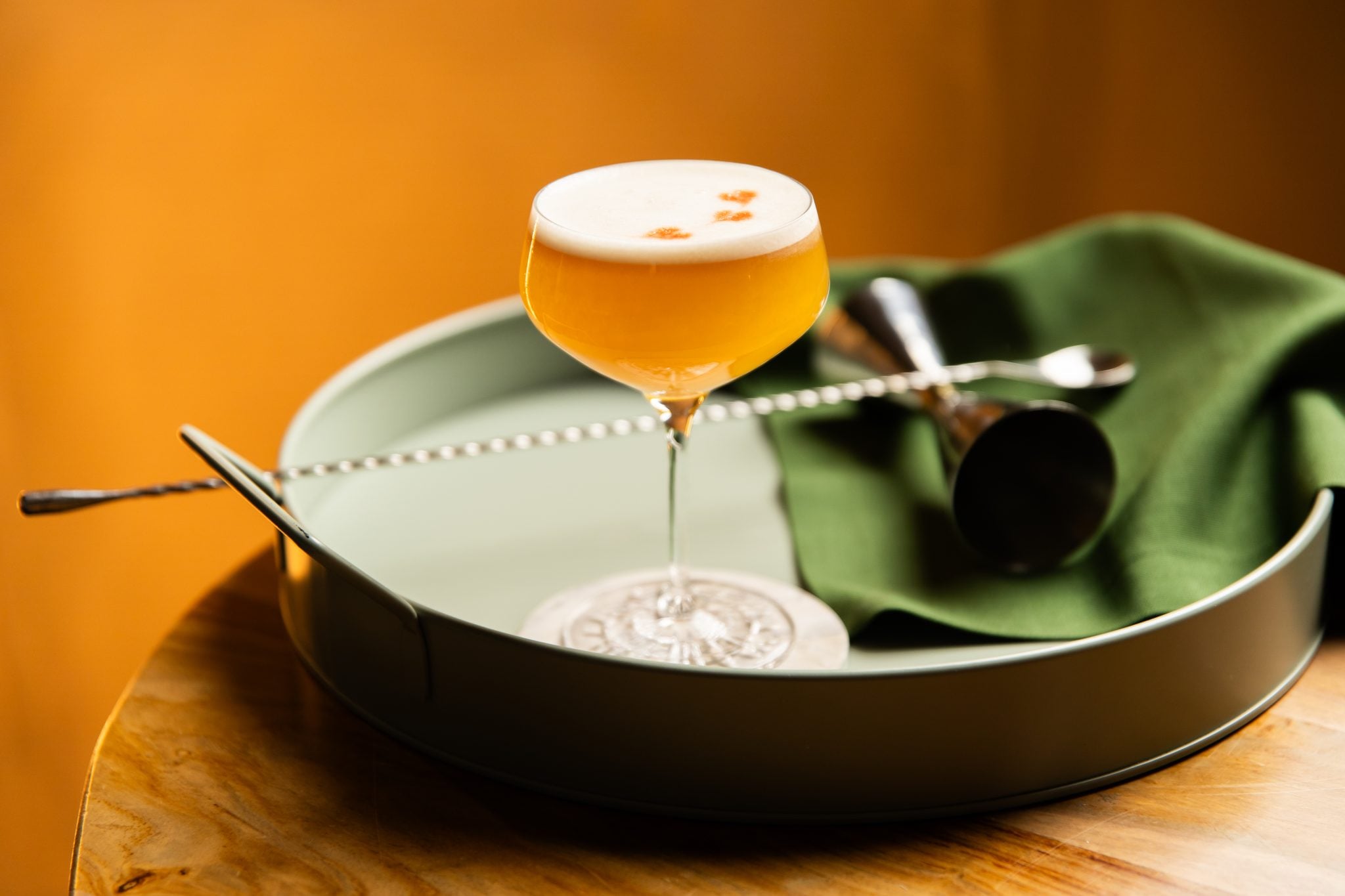 A side shot of a Japanese Sour cocktail in a coupe glass on a turquoise tray placed on a wooden surface surrounded by a bar spoon, a jigger and a green cloth