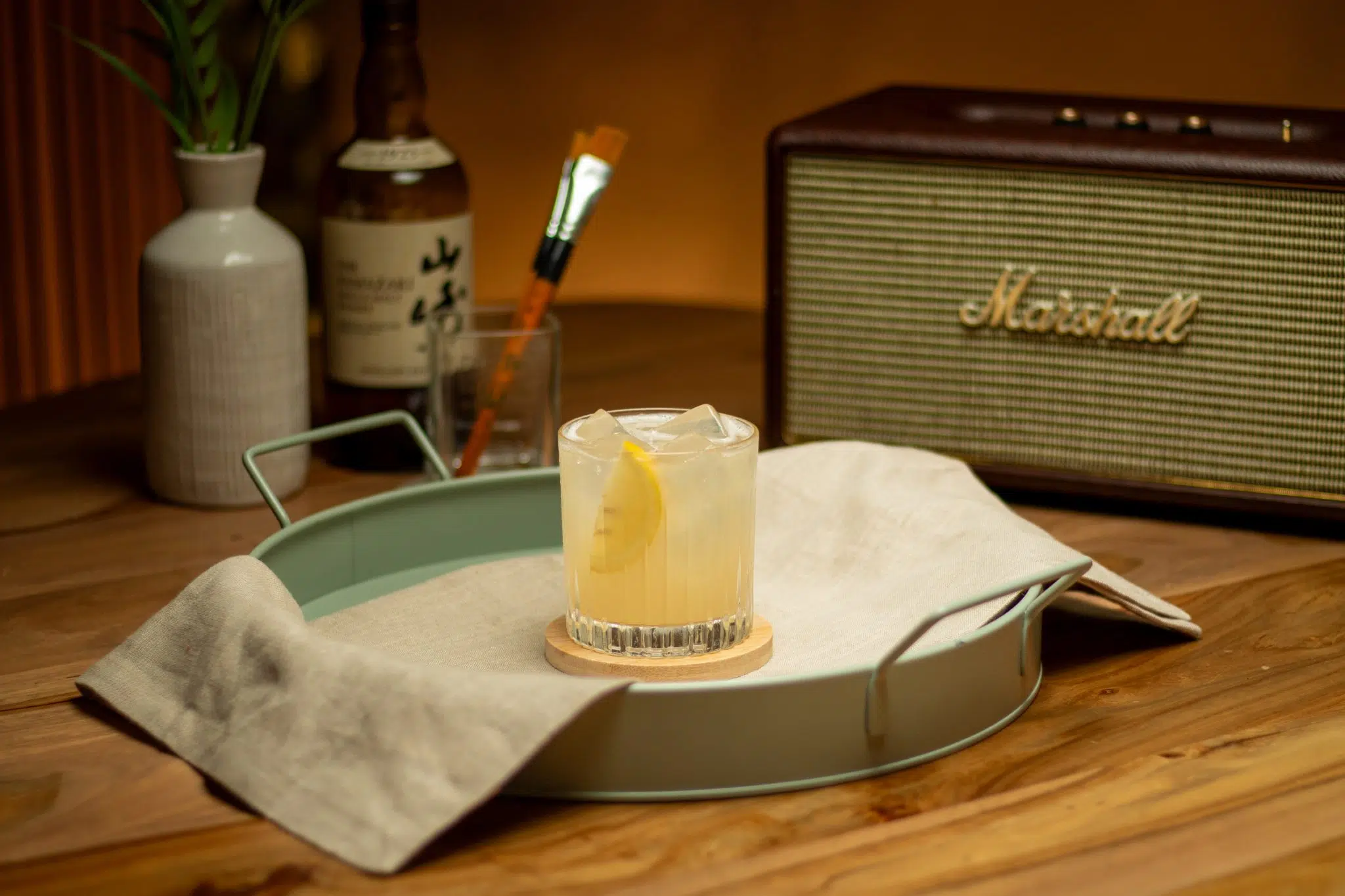 A side shot of a Japanese Yuzu Whisky Sour cocktail in an Old Fashioned glass on a turquoise tray on a wooden surface surrounded by a speaker, a Yuzu Whisky bottle and a white flower vase