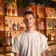 Joshua Miller smiling and standing in front of a bar with shelves of various bottles.