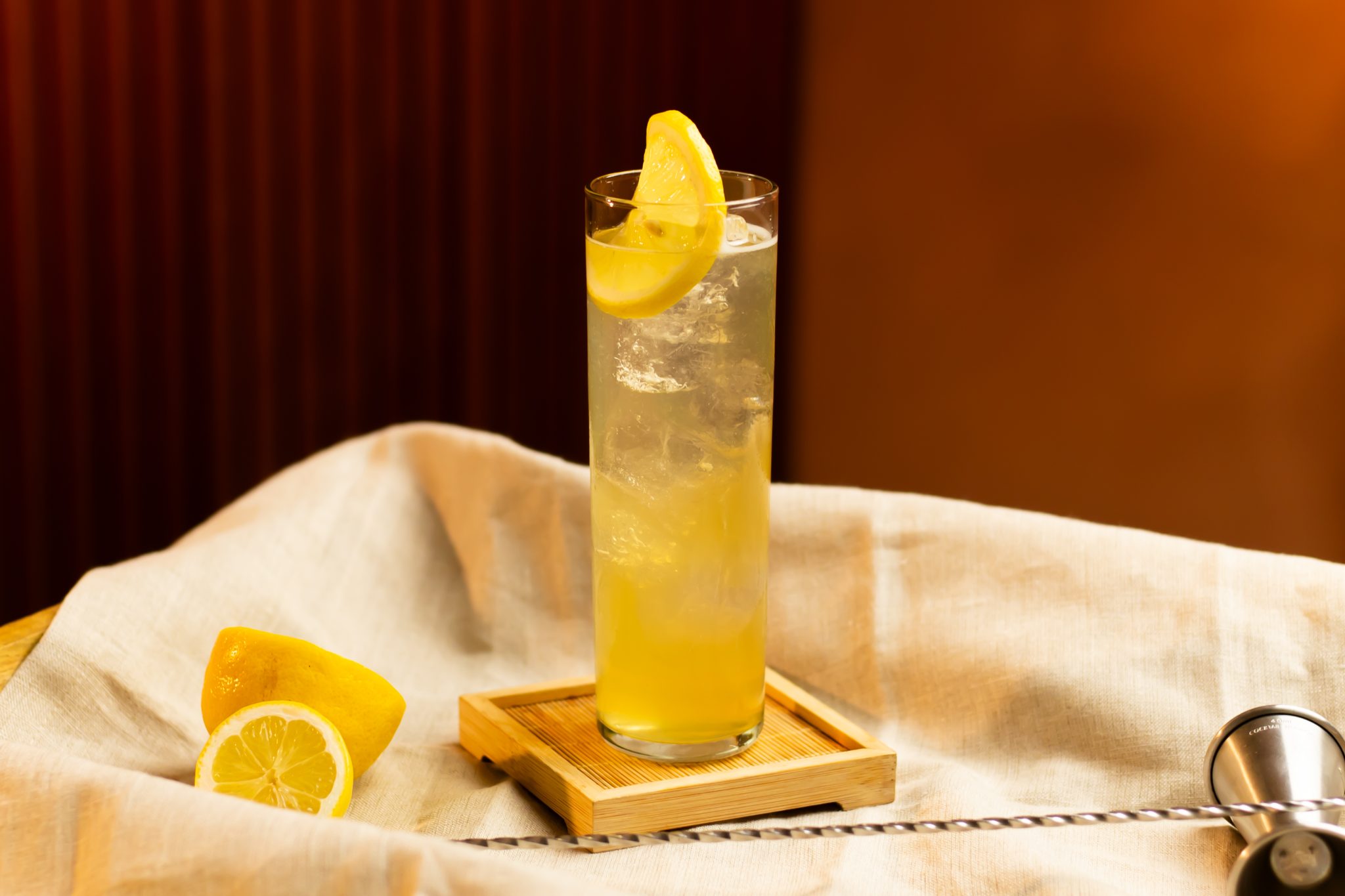 A side shot of a Juan Collins cocktail in a collins glass on a wooden coaster placed on a white cloth surrounded by two lemons, a bar spoon and a jigger, in front of a orange background