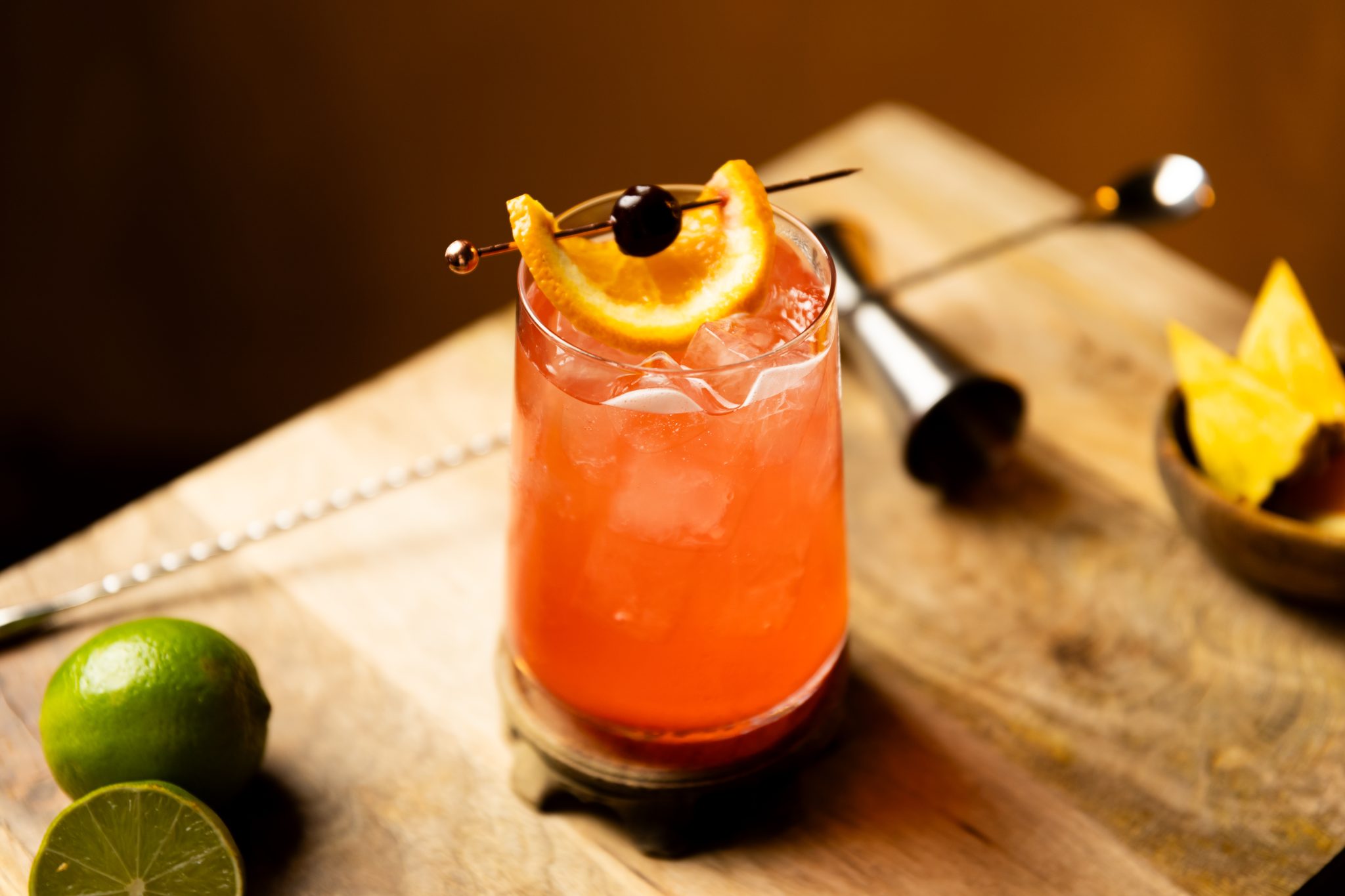 A side shot of a Jungle Juice cocktail in a highball glass on a wooden coaster and board surrounded by a jigger, a bar spoon, two limes and a bowl with pineapple pieces