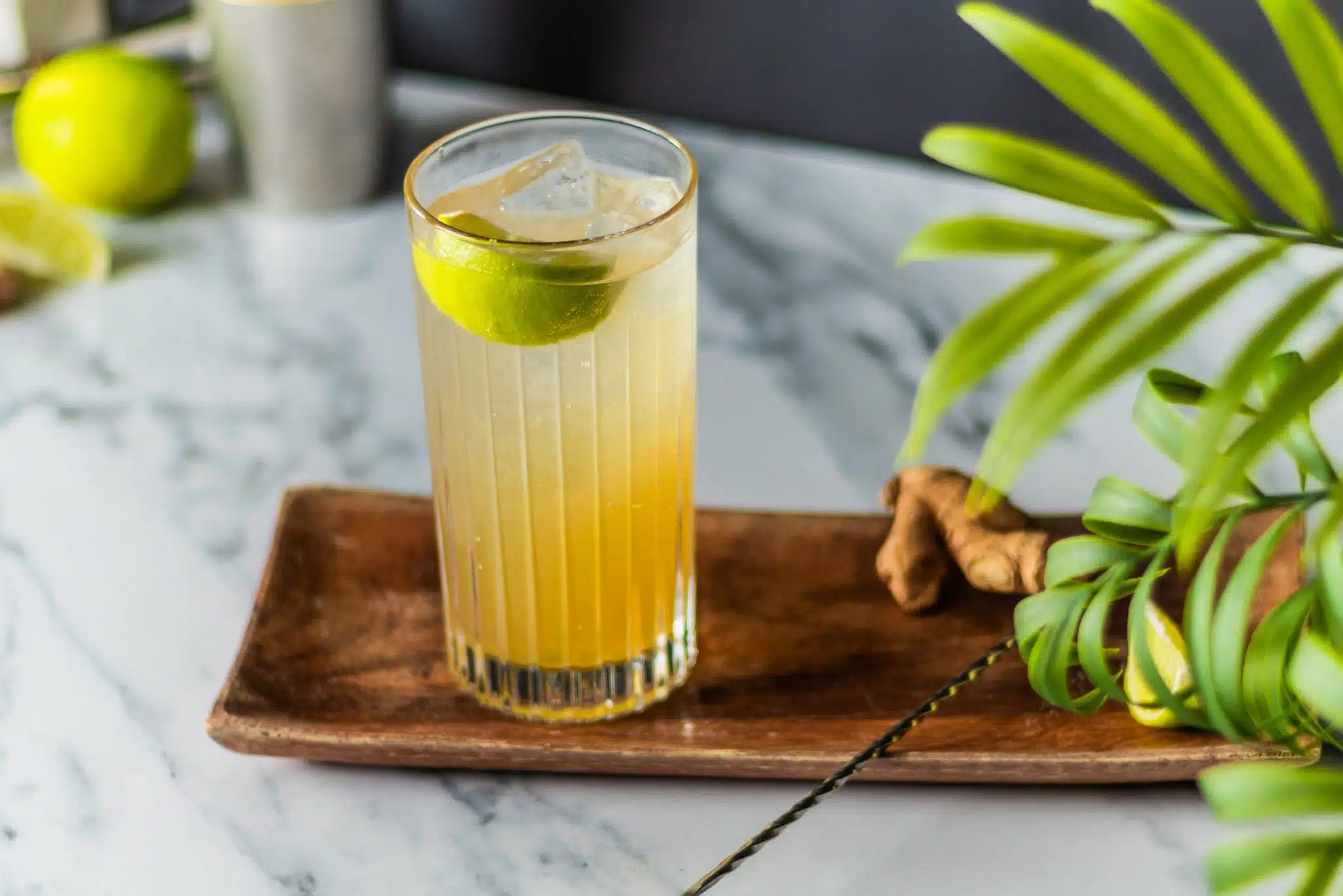A side shot of a Kentucky Mule cocktail in a highball cocktail with a ginger pieces and a bar spoon on a wooden tray placed on a white marmol table with a shaker and a lime on the background.