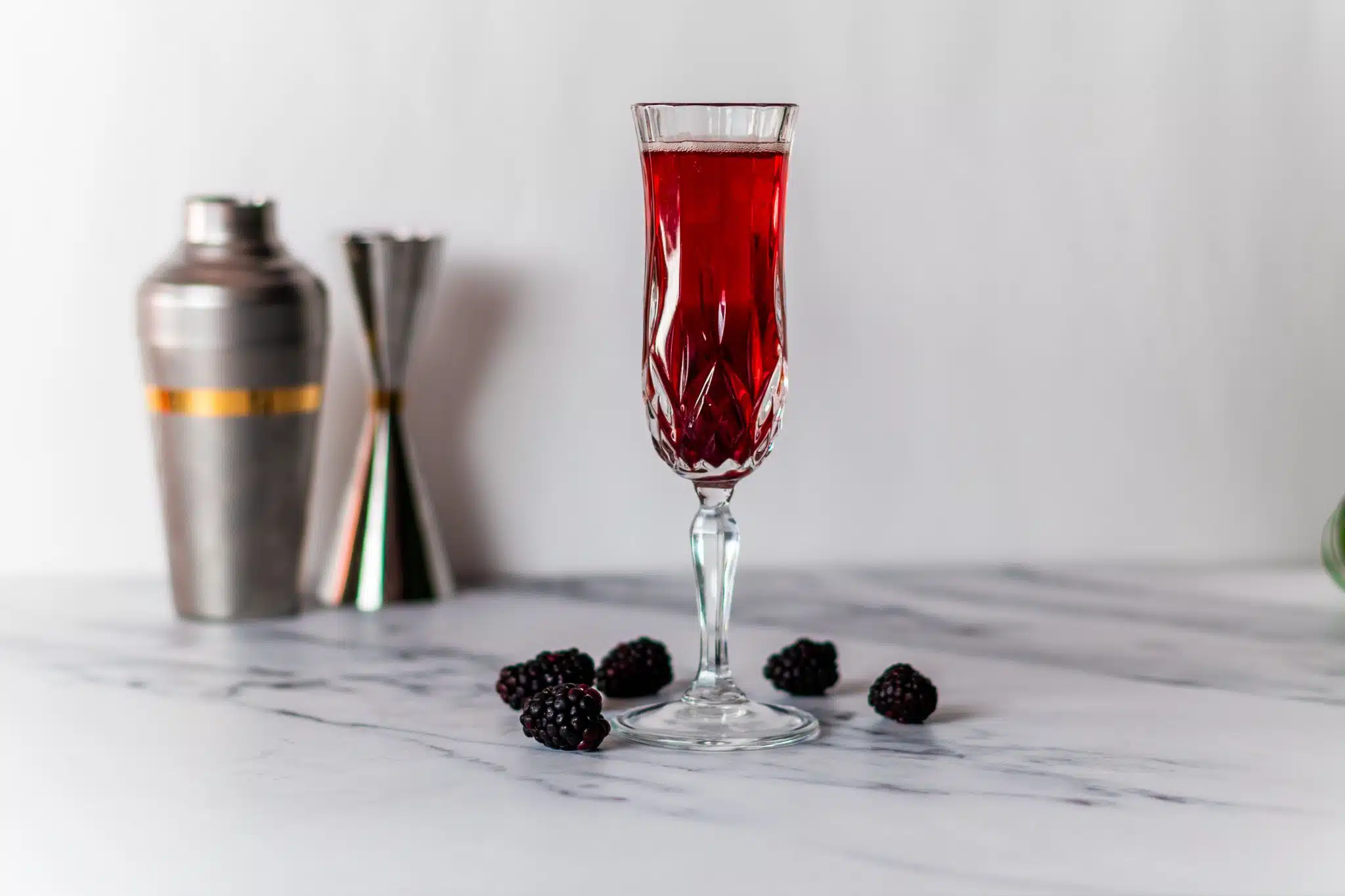 A side shot of a Kir Royal cocktail in a champagne flute on a white marmol table surrounded by berries and a shaker and a jigger on the background