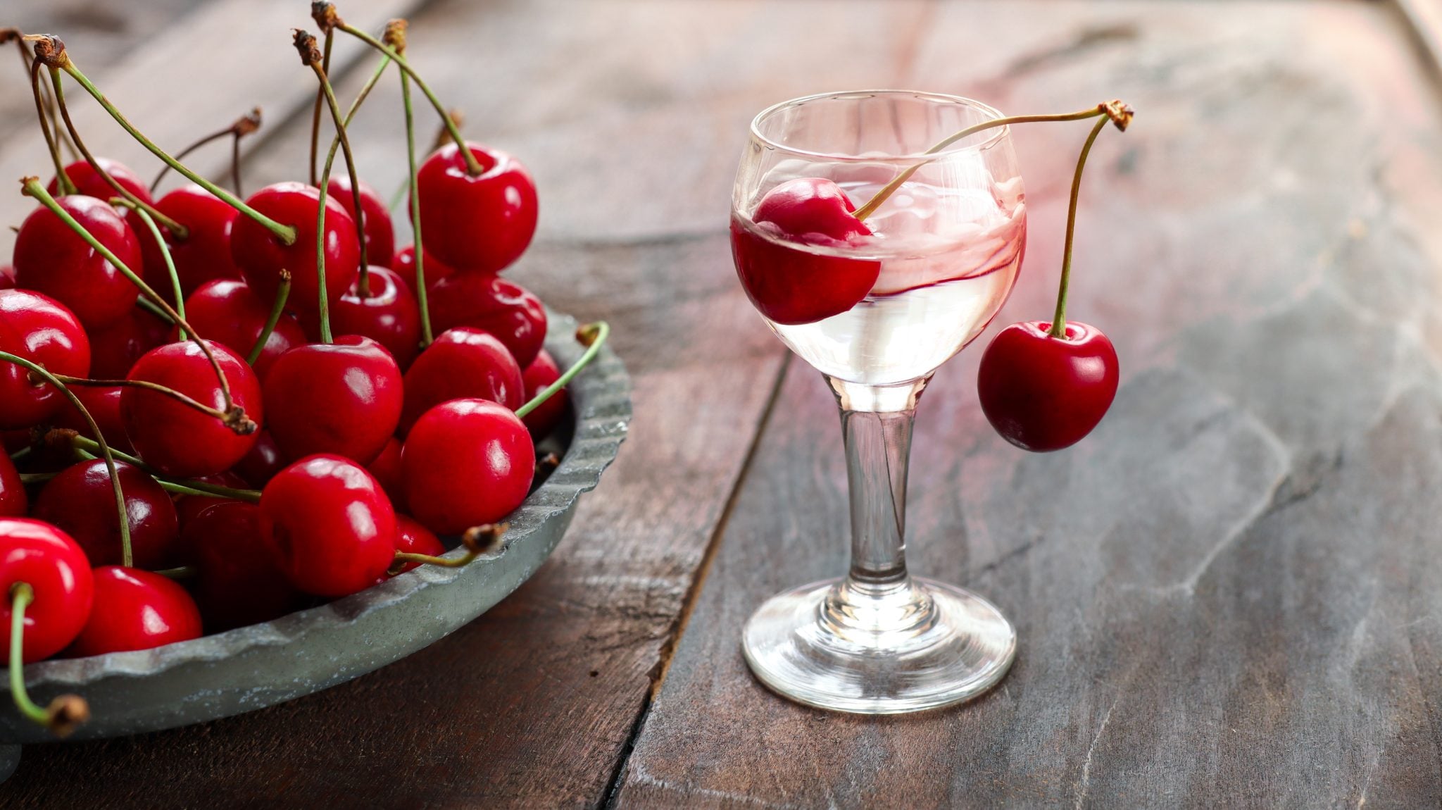A Kirschwasser shot garnish with two cherries on a wooden surface with a plate full with more cherries