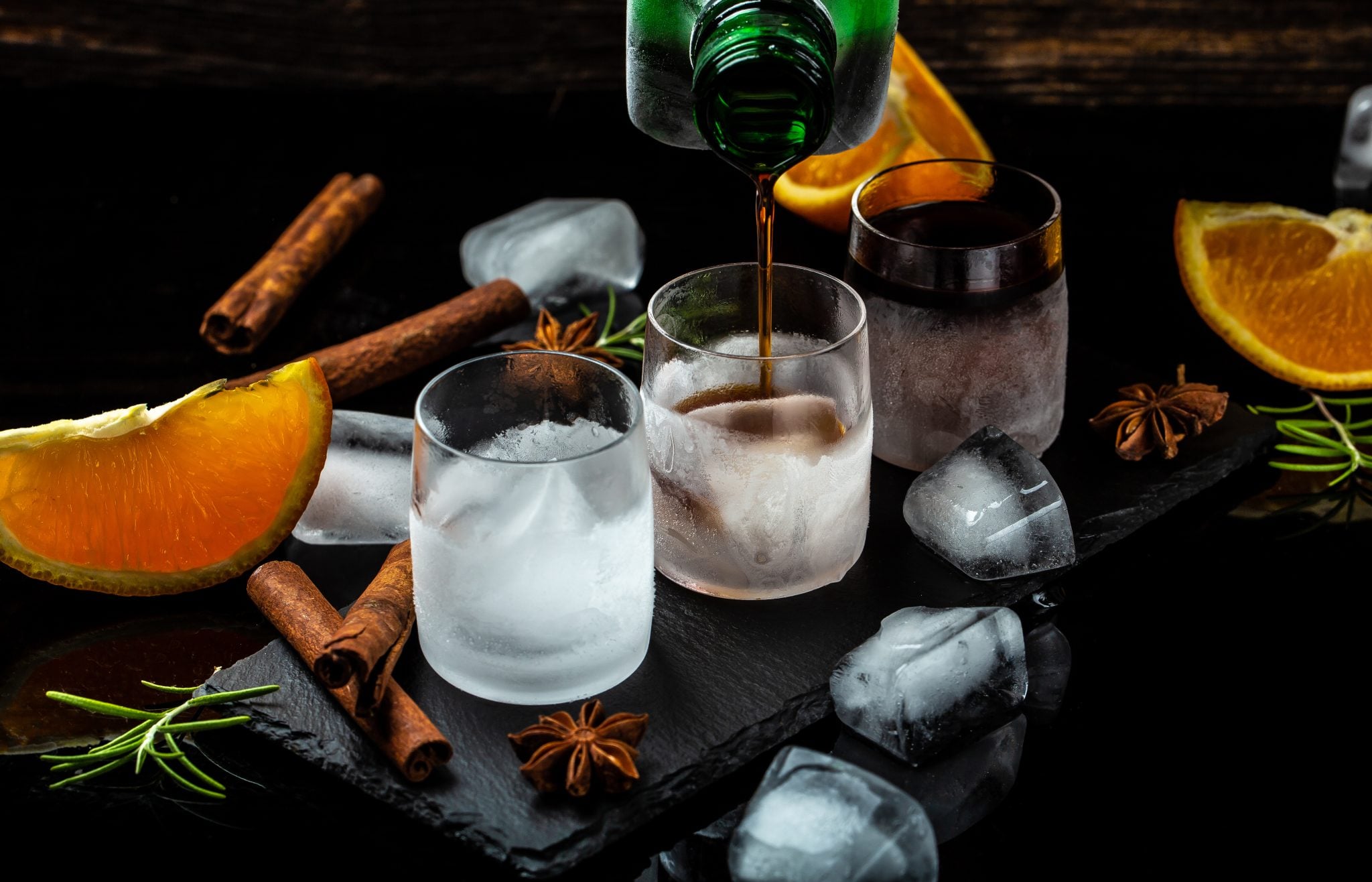 Kräuterlikör poured into chilled shot glasses on a black surface surrounded by orange wedges, cinnamon sticks, and ice cubes
