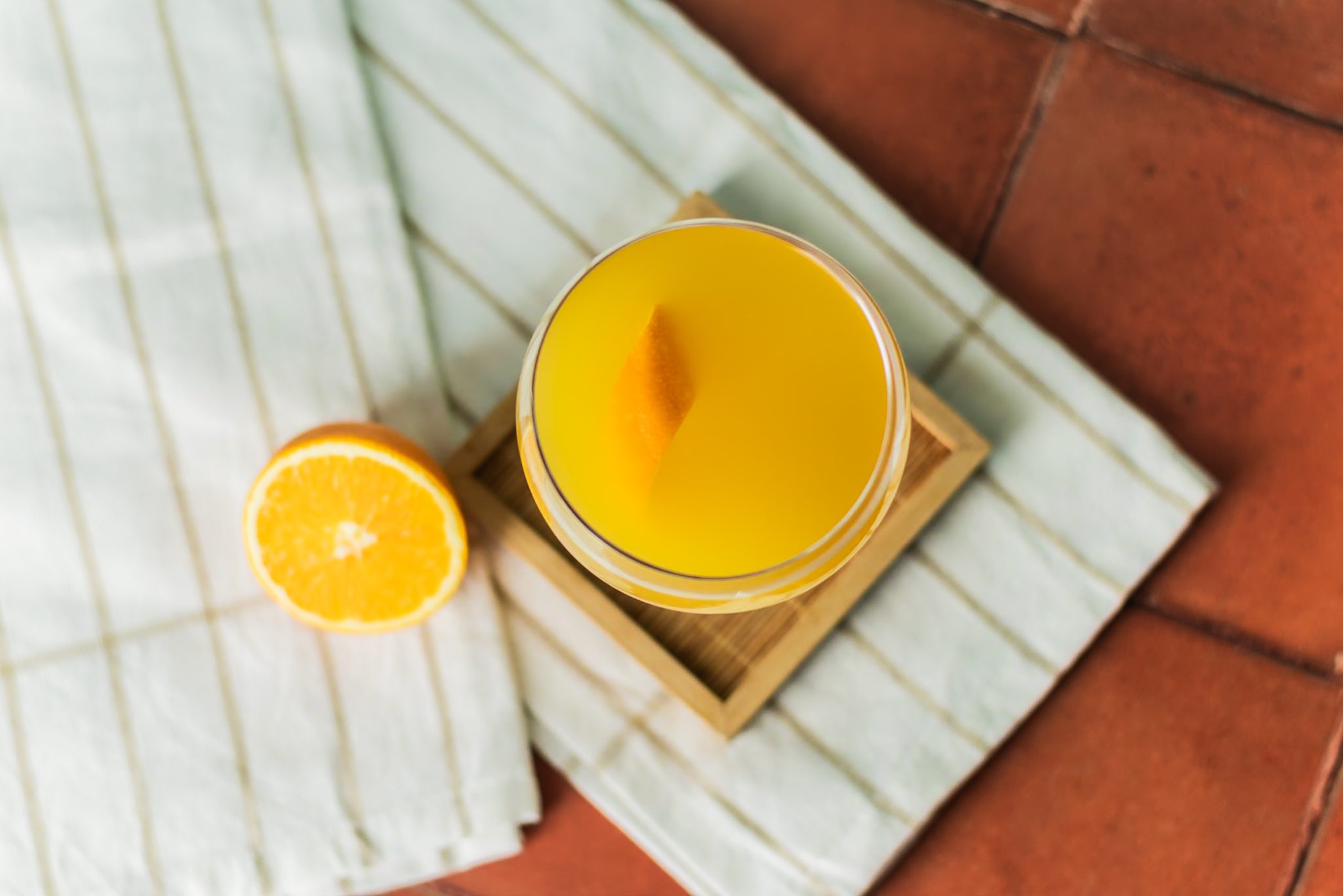 A La Valencia cocktail, shot form above, in a cocktail glass on a coaster placed on a striped white cloth on a tiled surface with an orange around.