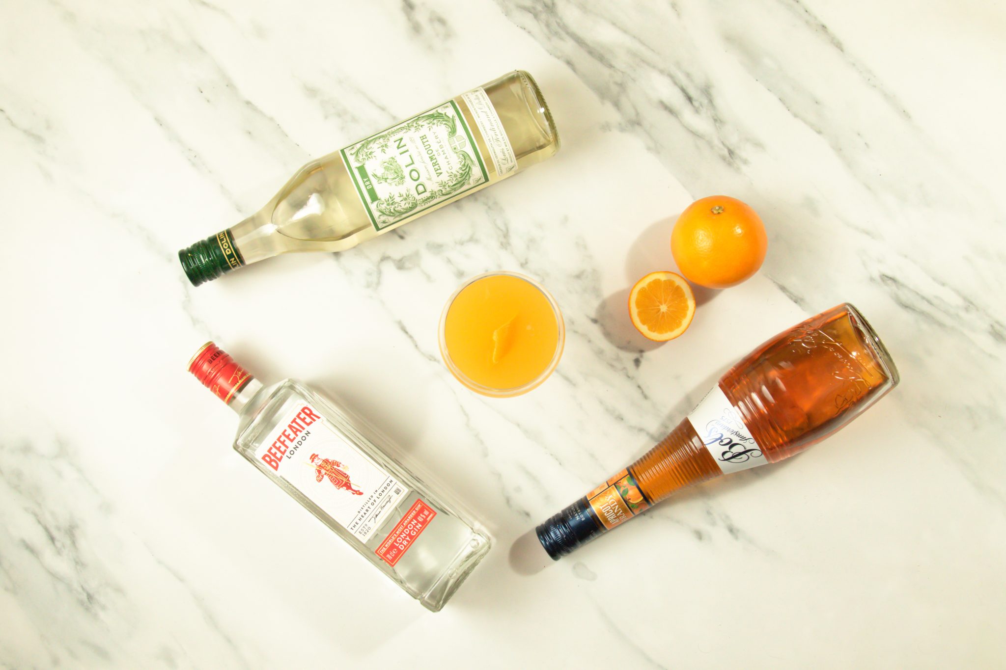 A La Valencia cocktail, shot from above, in a cocktail glass on white marmol table surrounded by a Gin bottle, an apricot Brandy bottle, a dry Vermouth bottle and two oranges.