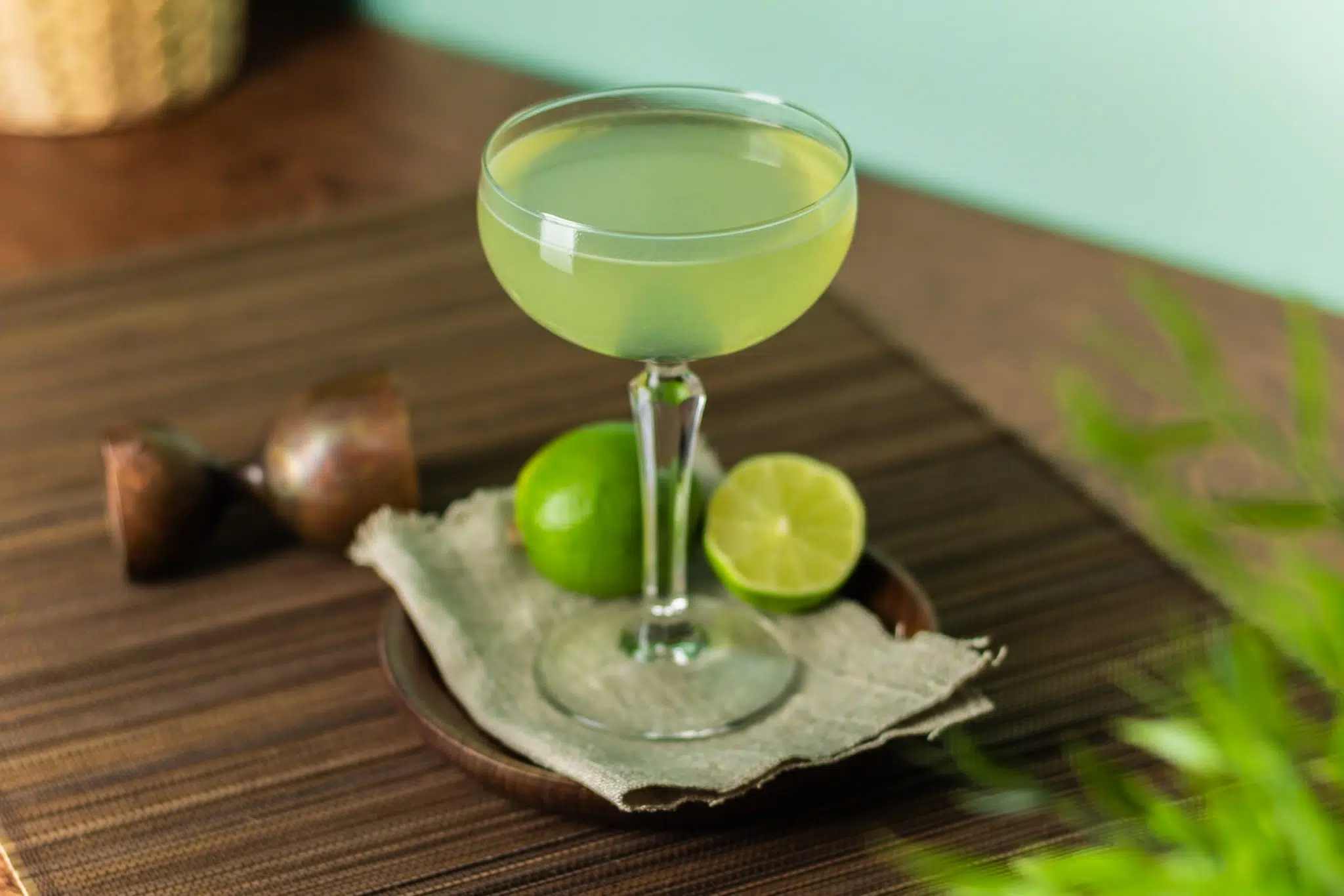 A Last Word cocktail, shot from above, in a cocktail glass on a smal plate placed on a brown placemat with a jigger, two limes, a white cloth around.