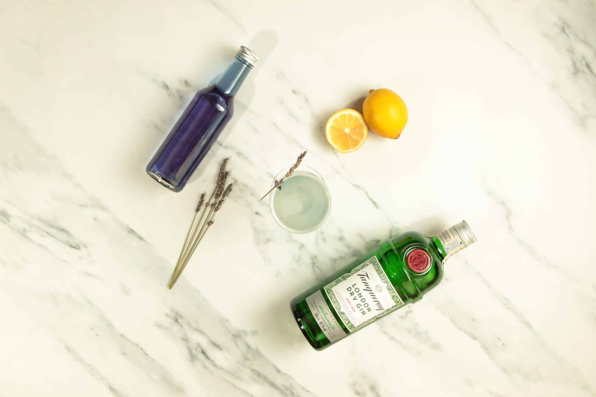 A Lavender Martini cocktail, shot from above, on a white marmol table with a gin bottle, a lavender simple syrup bottle, lavender sprig and two lemons around.