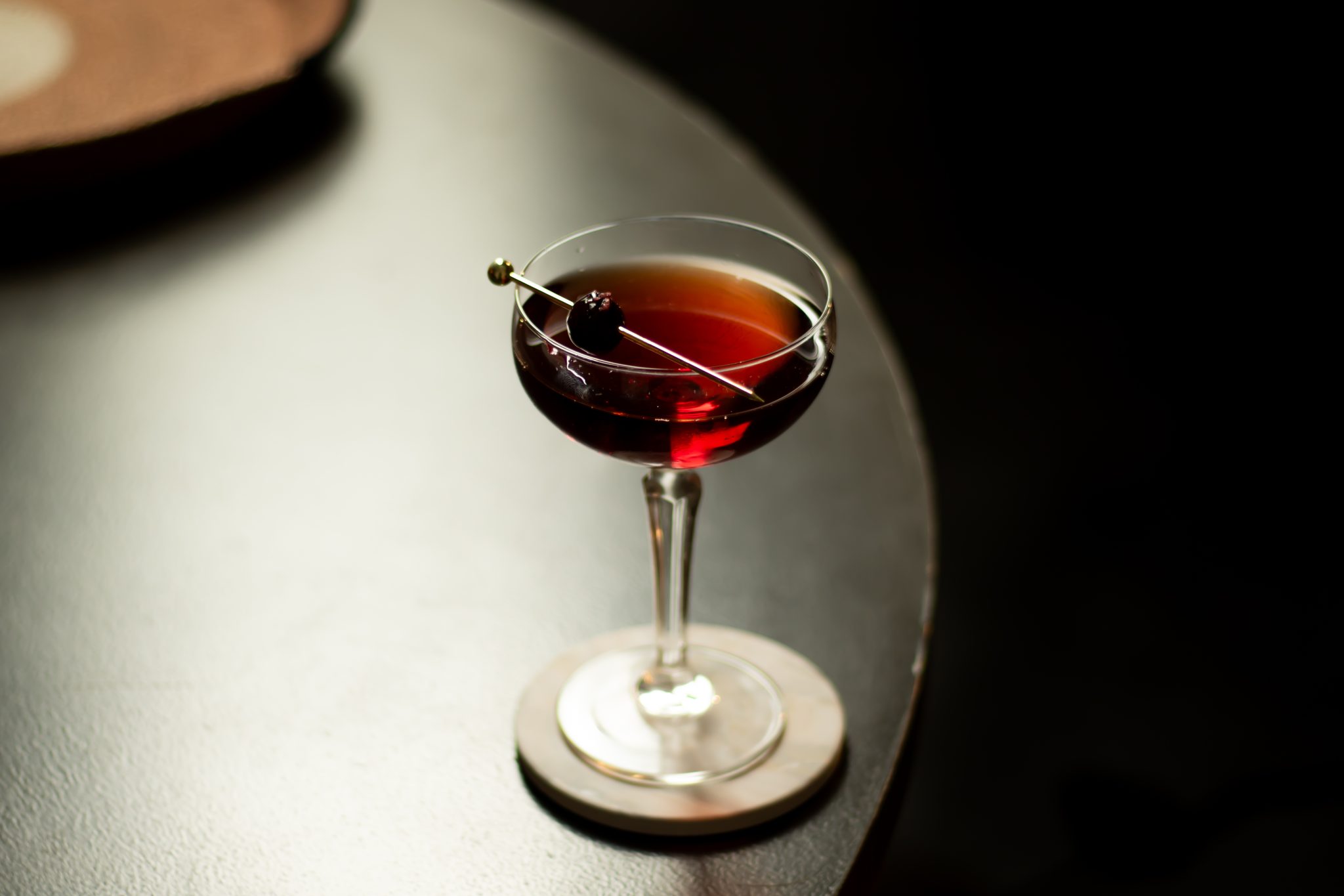 A side shot a Left Hand cocktail in a coupe glass on a white coaster on a black table in front of a black background