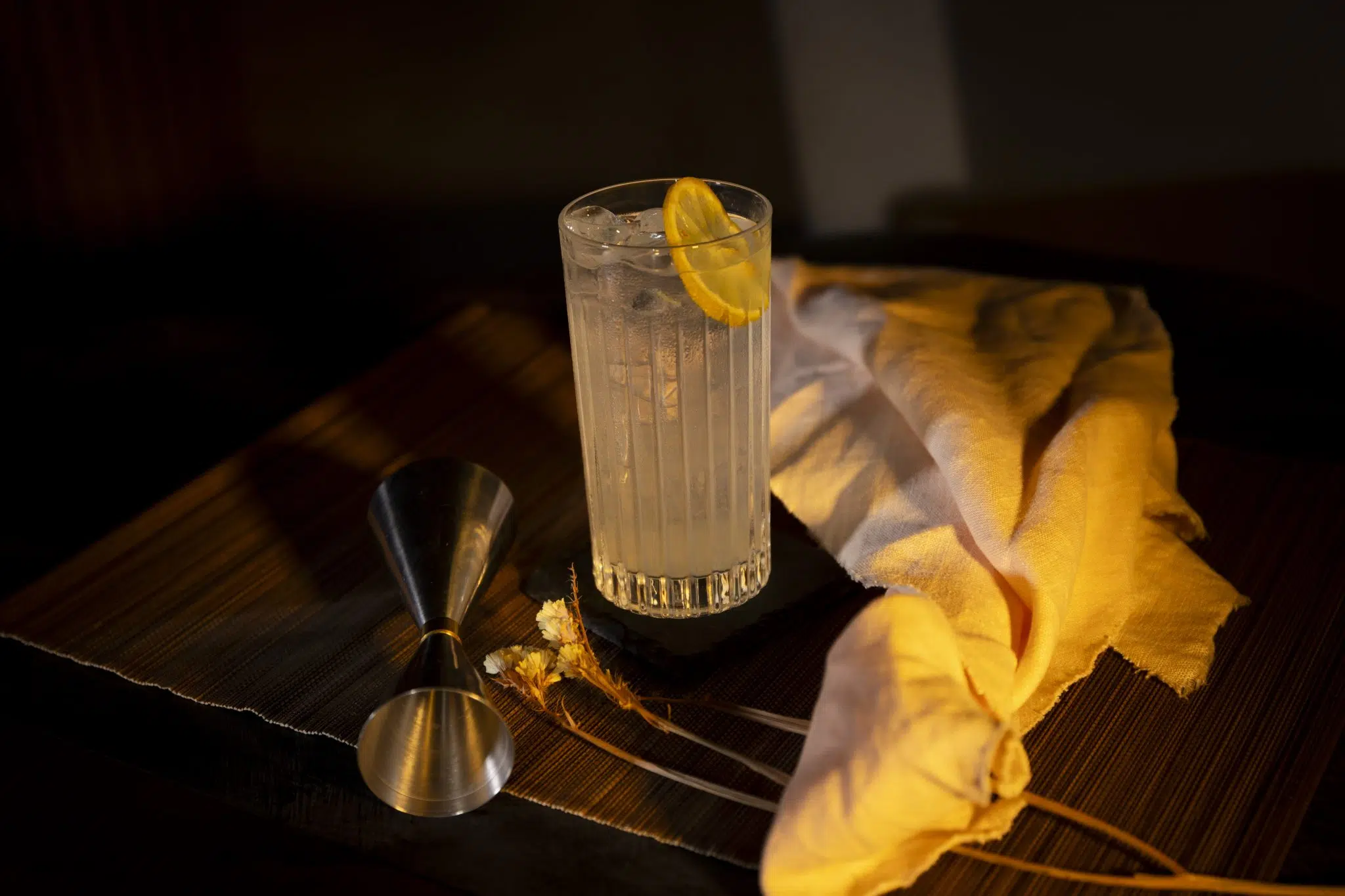 A side shot of a Lemon Drop Highball cocktail in a highball glass on a brown placemat surrounded by a jigger, a flower sprig and a white cloth