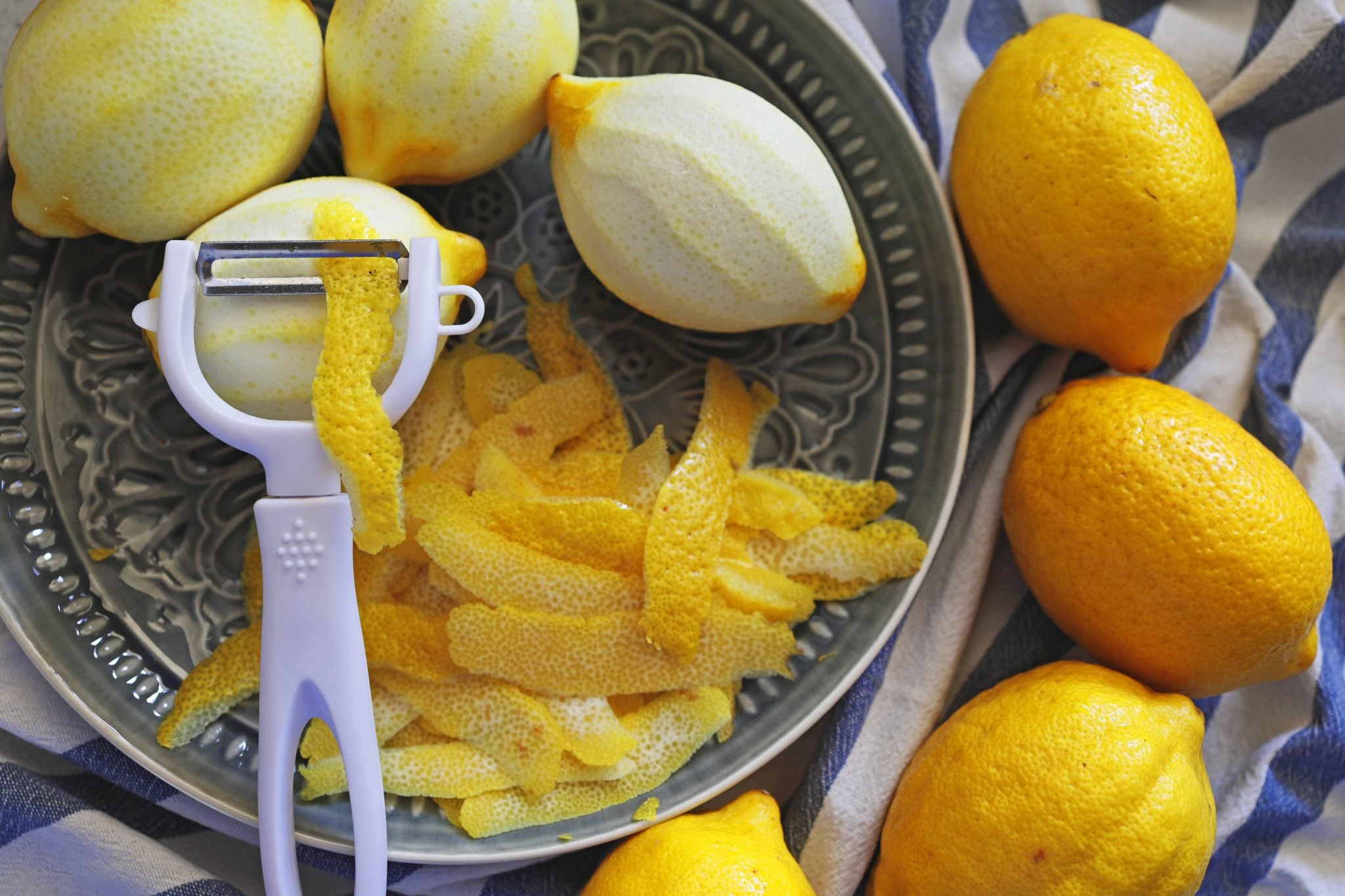 A shot from above of three peeled lemons, four lemons, a peeler, and many peels on a plate