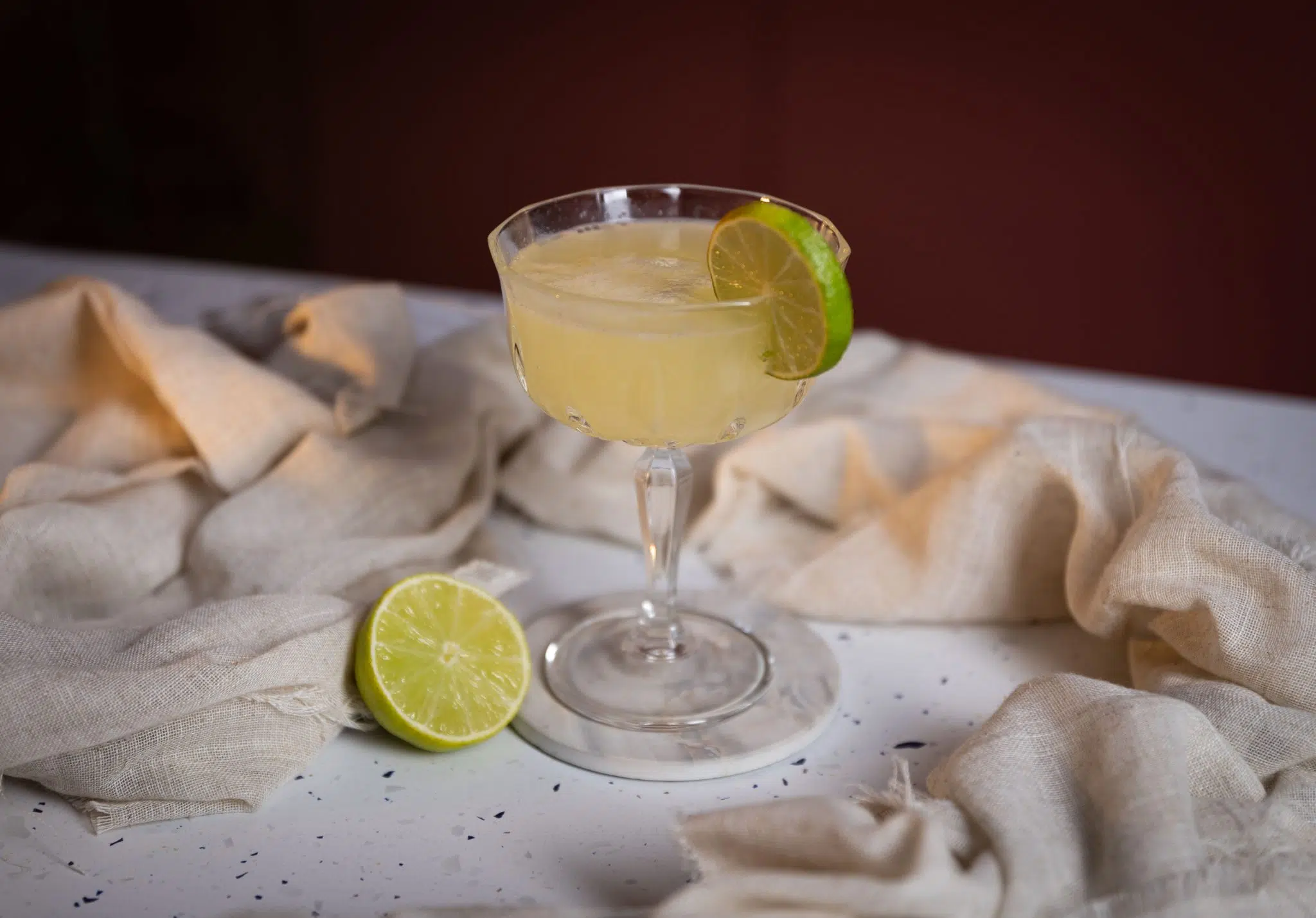 A side shot of a Lime Sherbet Punch cocktail in a coupe glass on a white coaster and tablecloth surrounded by half a lime, and a beige cloth
