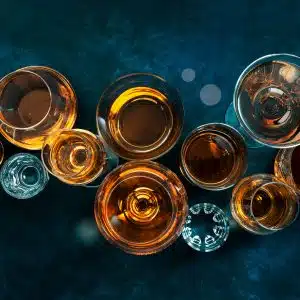 11 glasses filled with liqueurs on a dark smokey blue surface.