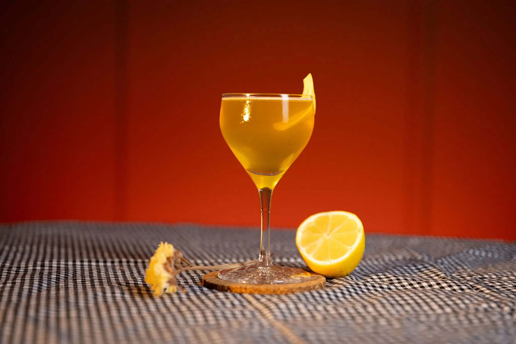 A side shot of a Loose Talk cocktail in a coupe glass on a cork coaster placed on a black and white tablecloth surrounded by half a lemon and a flower sprig