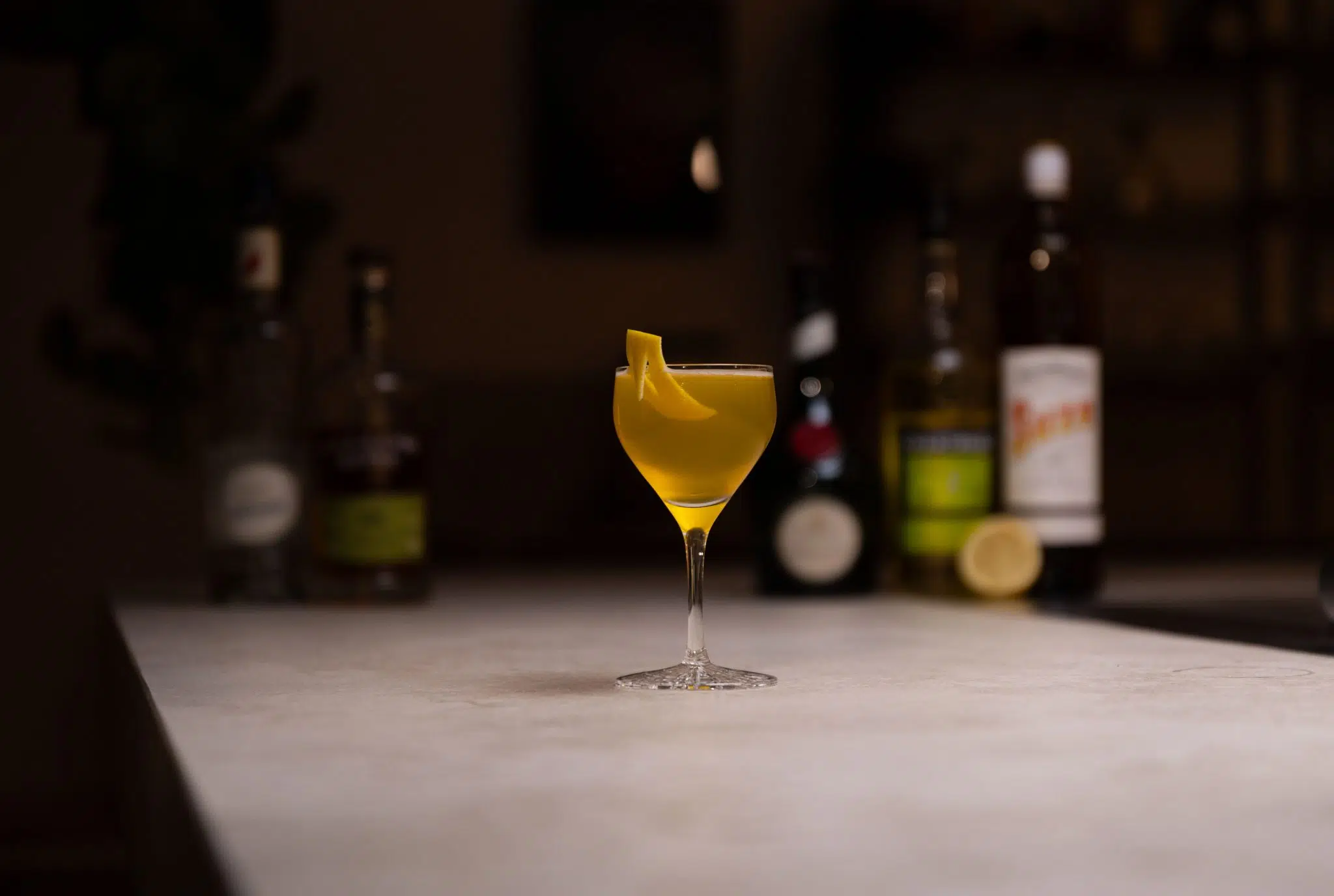 Rye whiskey, gentian liqueur, yellow Chartreuse, lemon juice, lime juice, maraschino liqueur and Benedictine liqueur laid out on a white bar table