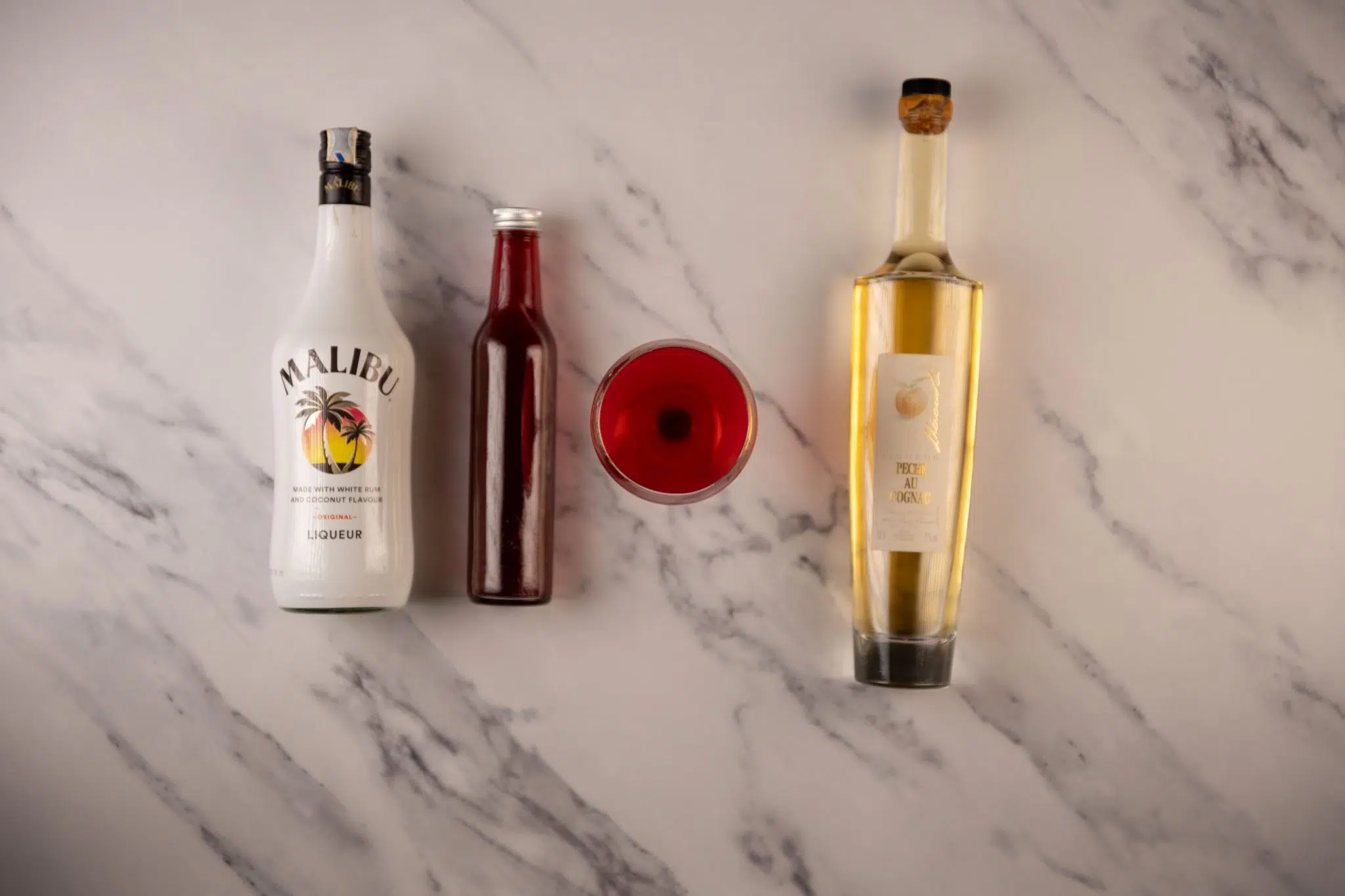 A Love Martini cocktail, shot from above, in a martini glass on a white marmol surface surrounded by Peach Schnapps, cranberry juice, and coconut rum