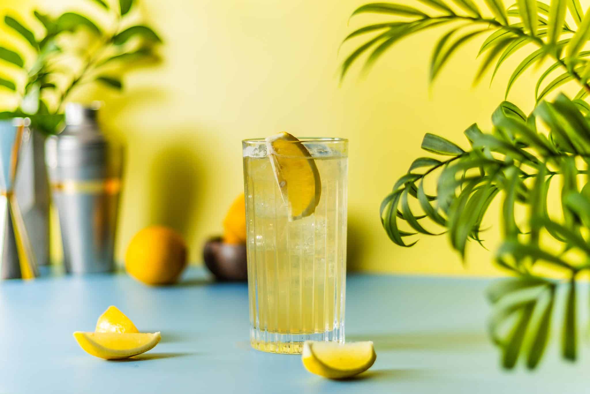 A side shot of a Lynchburg Lemonade cocktail in a highball glass surrounded by lemon wedges and plant leaves on a blue table, with some cocktail tools behind placed in front of a yellow wall.