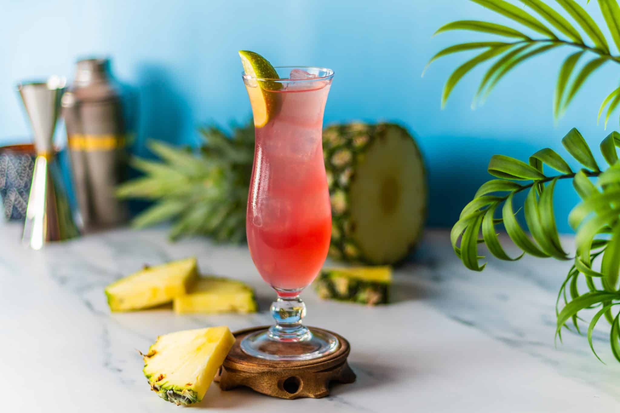 A side shot of a Malibu Bay Breeze cocktail in a hurricane glass on a wooden coaster placed on a white marmol table with pineapple wedges around.