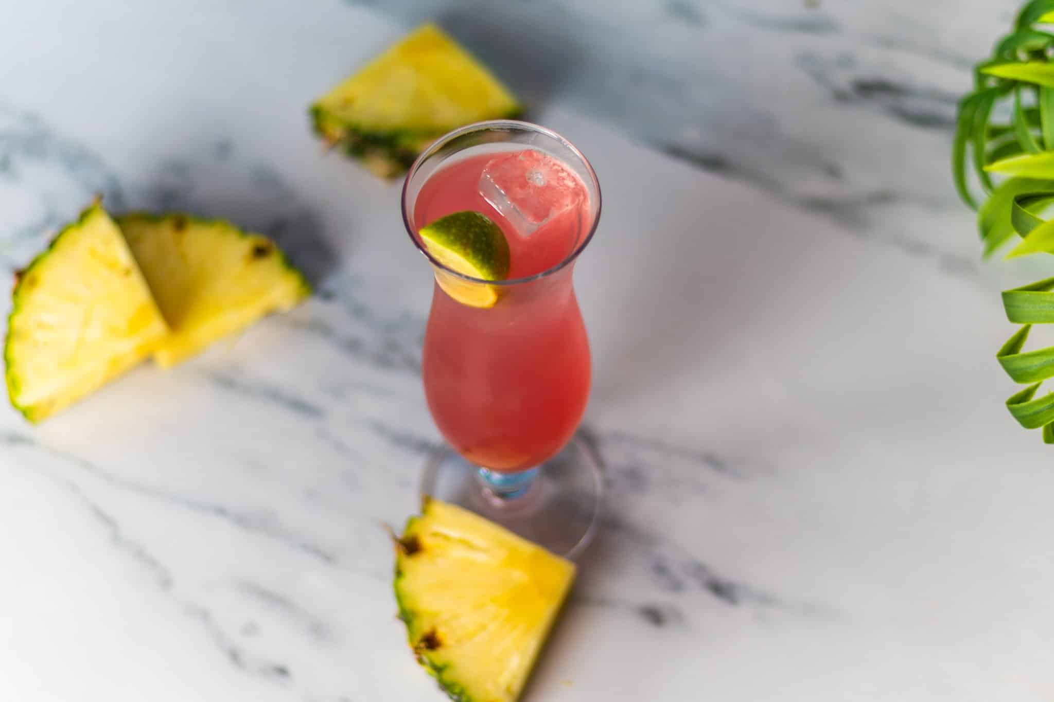 A Malibu Bay Breeze cocktail, shot from above, in a hurricane glass on a white marmol table surrounded by pineapple wedges.