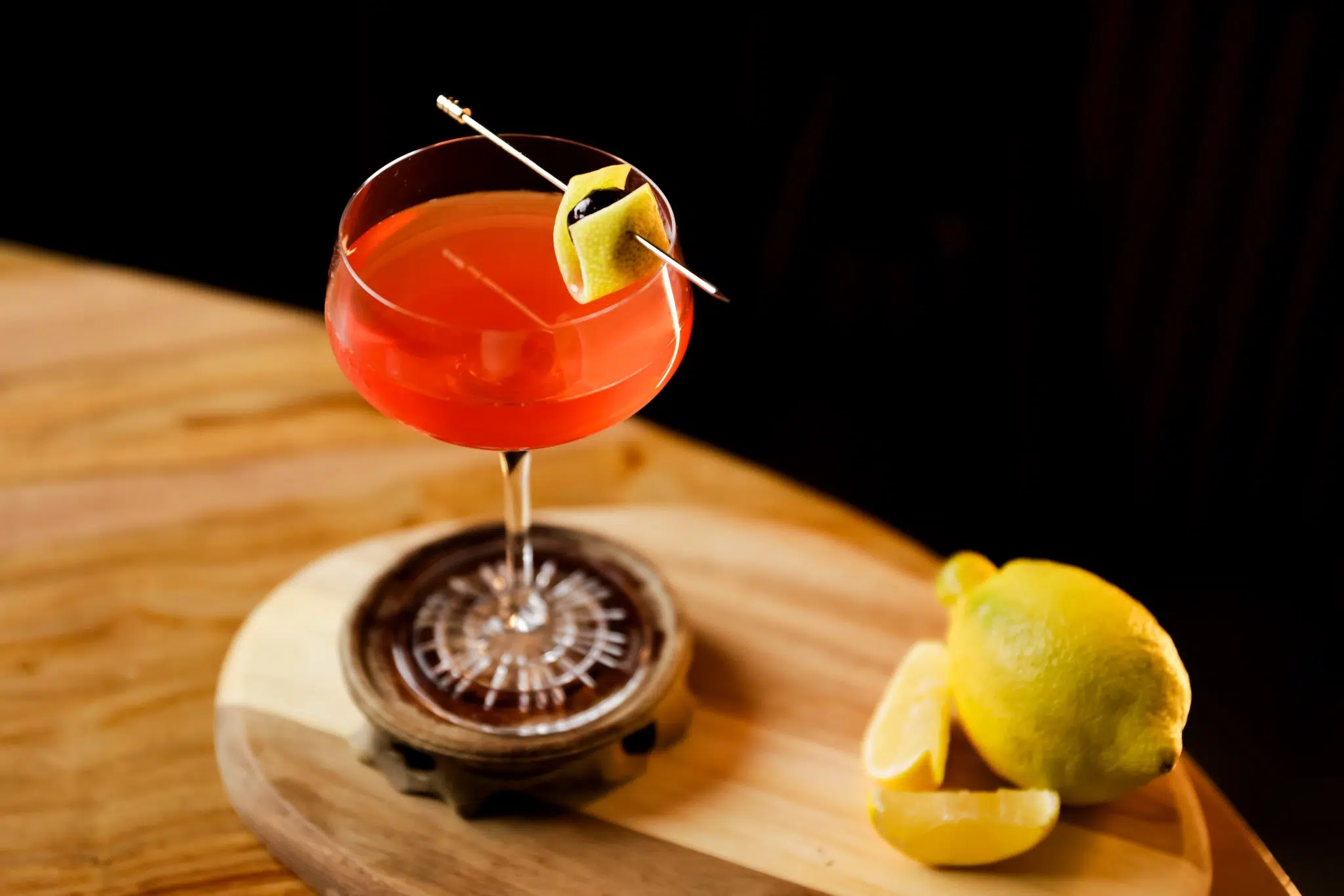 A side shot of a Man O' War cocktail in a coupe glass on a wooden coaster, board and table with a lemon and a lemon zest in front of a black background