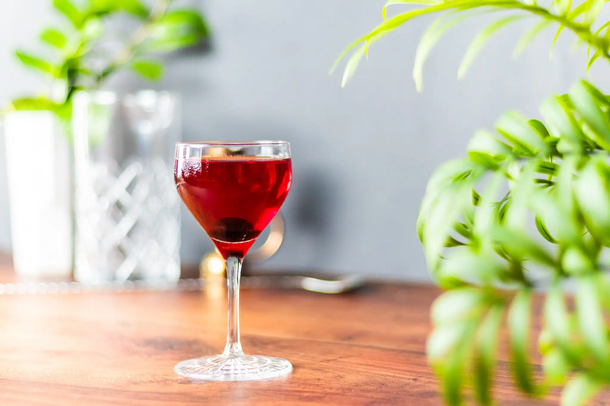 A side shot of a Manhattan cocktail in a cocktail glass on a brown wooden table with a plant on the side and a bar spoon and mixing glass on the background.