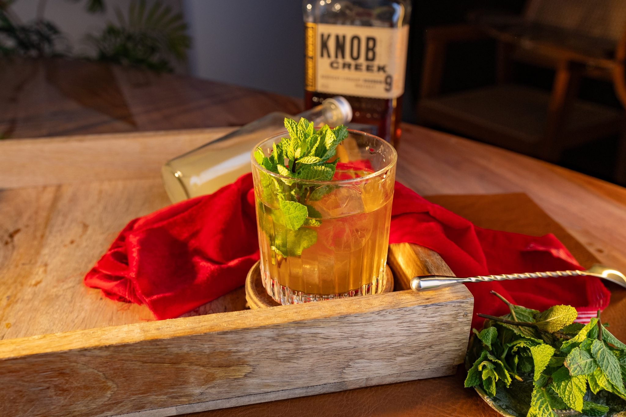 A side shot of a Maple bourbon Smash cocktail in an old fashioned glass on a wooden coaster and tray surrounded by a red cloth, a bourbon bottle, a maple syrup bottle, a bar spoon and mint leaves